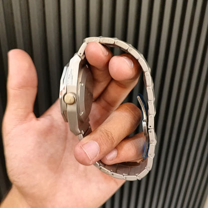 A close-up of a person's hand holding a silver-toned metal watch with a matching link bracelet, showing the side profile of the watch case and crown.